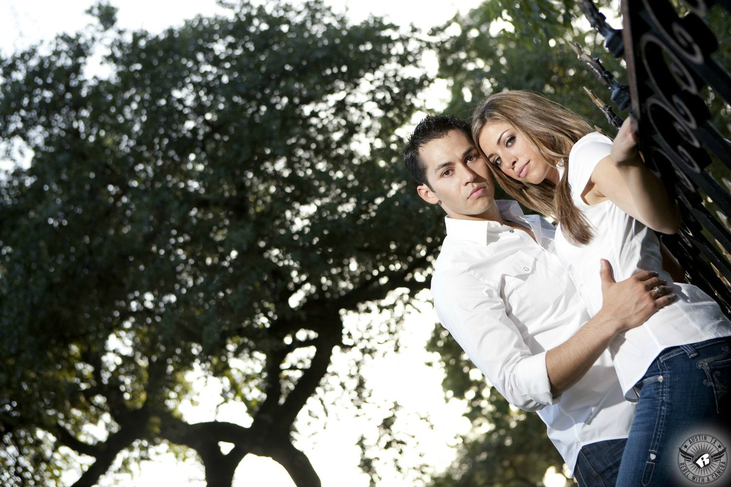 Strawberry blond Latino girl in white short sleeve top and blue jeans is held at waist by dark spiky haired guy wearing white shirt unbuttoned at the top and dark blue jeans lean on black rod iron fence at the Texas Capital with the bright sky and out of focus trees in this edgy engagement picture in Austin.